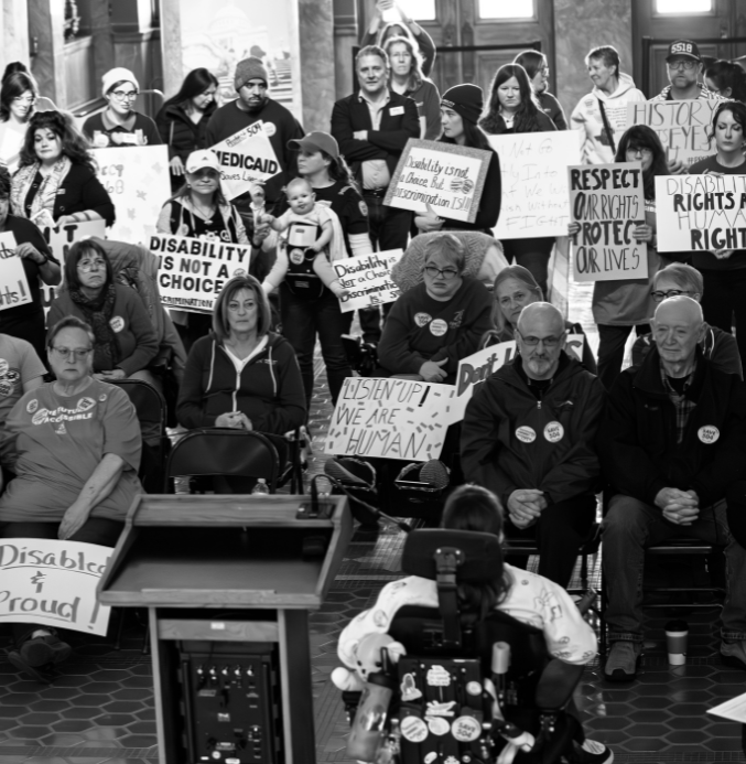 Crowd of people holding various signs in support of Disability Rights while listening to a speaker who is a wheelchair using sitting next to the podium.
