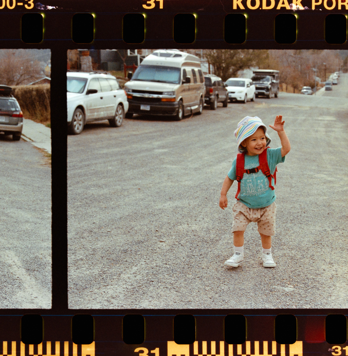 35mm film scanned showing sprocket holes, a child walking down a dirt road wearing a backpack and waving to someone outside the image.