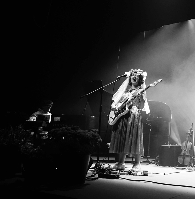 Black and white image of a woman playing guitar and singing loudly into a microphone. Dramatic lights, fog in background.